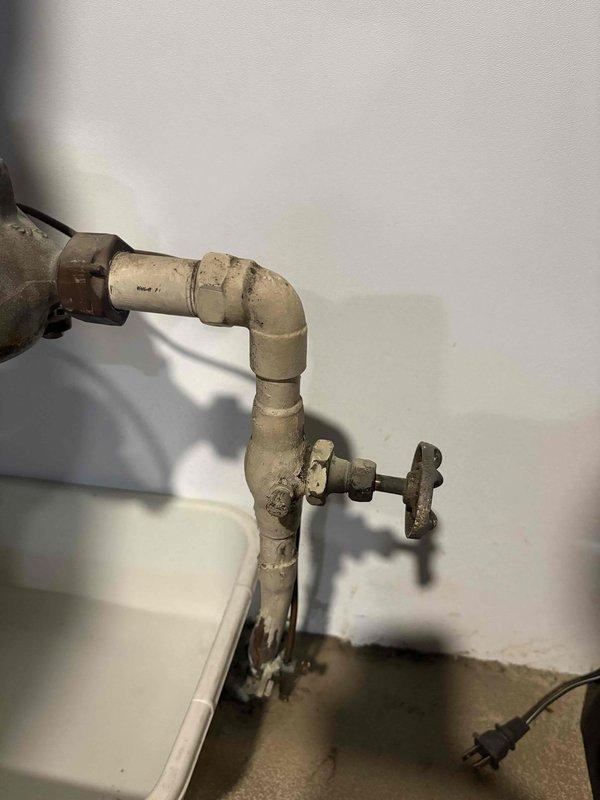 # Outdoor Spigot Leak Repair

Responded to a water leak at the basement shut-off valve serving an exterior hose spigot. Upon inspection, identified active leaking at the aged galvanized pipe assembly where the outdoor spigot line connects to the main water distribution system. The existing piping showed significant corrosion and weathering consistent with years of service, with visible oxidation at connection points and the shut-off valve mechanism.

Shut down water supply to the affected line and assessed the plumbing configuration, which consisted of a vertical feed pipe with a T-junction and cross-handle valve mounted against the basement wall. Water staining on the adjacent wall surface confirmed ongoing moisture intrusion from the compromised connection. 

Disassembled the leaking joint at the shut-off valve and evaluated the condition of the threaded connections. Cleaned pipe threads, applied appropriate thread sealant, and reassembled the connection with proper torque specificat