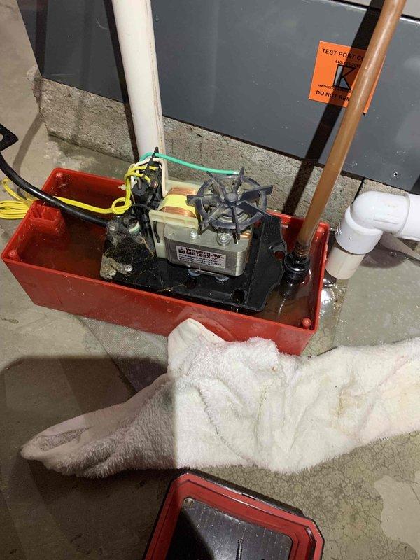 Responded to service call for leaking condensate pump on air handler system. Upon arrival, found active water leak from condensate pump assembly with moisture present on concrete floor surrounding red containment basin. Inspected pump components including motor housing, discharge piping, and connections. Placed absorbent materials to contain water and prevent further floor damage. Documented existing conditions with photographs showing pump assembly, connected piping, white collection container, and HVAC furnace installation. System evaluated for proper drainage function and pump operation to identify source of leak and determine necessary repairs.