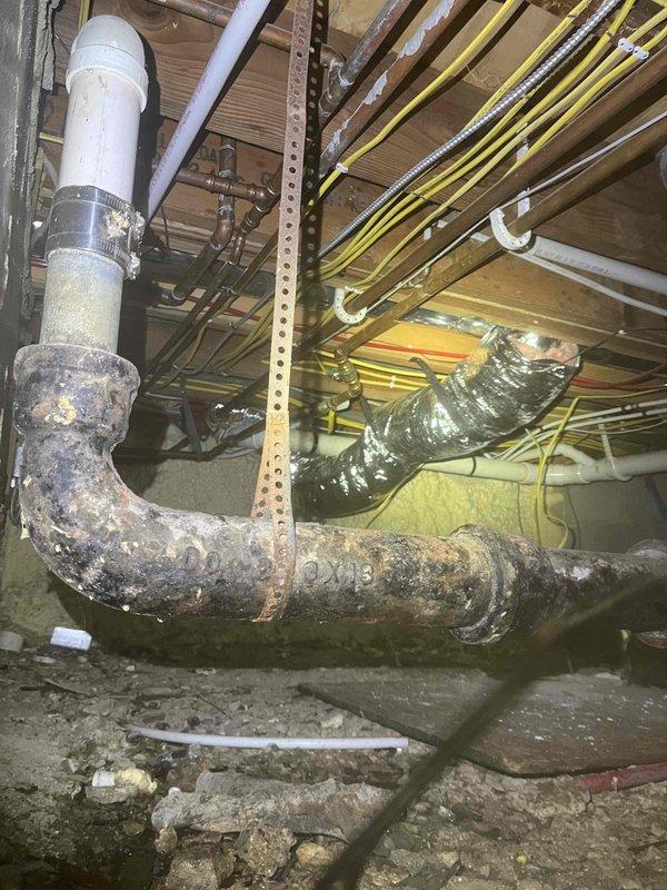 # Plumbing Service Completed

Responded to service call for gurgling sounds occurring in kitchen sinks during water usage. Upon investigation, accessed crawl space beneath the building to inspect the drainage system. Discovered severely corroded drain pipes showing extensive rust, surface degradation, and material deterioration on the main waste line. The corroded metal piping exhibited significant pitting and flaking, particularly on the horizontal drain run connecting to the kitchen sink system.

Documented the condition of the deteriorating pipe infrastructure, noting the transition point where newer white PVC piping connects to the older corroded metal sections. The severe corrosion on the main drain line was identified as the likely cause of restricted water flow and improper venting, resulting in the gurgling sounds reported by the customer. The deteriorated pipe surface and internal buildup can create air pressure imbalances within the drain system, producing characteristic gurg