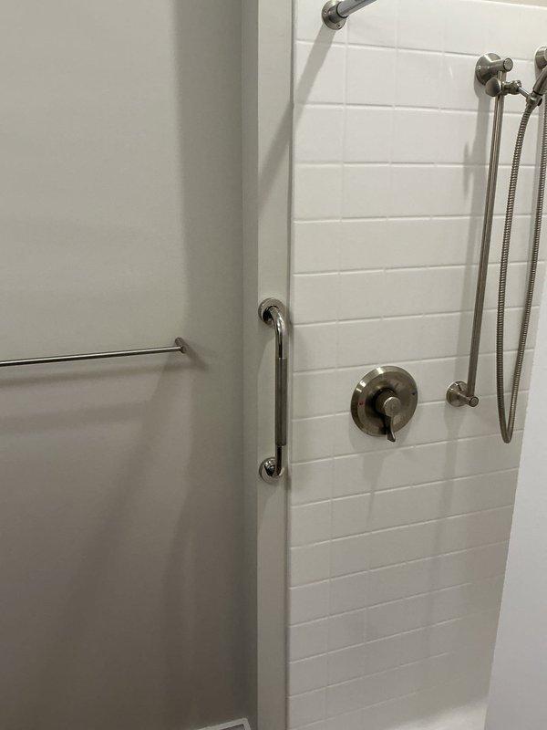 Evaluated existing shower supply line configuration and assessed feasibility of relocating water lines from attic space to accommodate revised bathroom layout per new construction plan. Inspected accessible bathroom installation featuring white subway tile surround, thermostatic mixing valve, handheld shower wand with slide bar, fixed showerhead, and multiple ADA-compliant grab bars in brushed nickel finish.