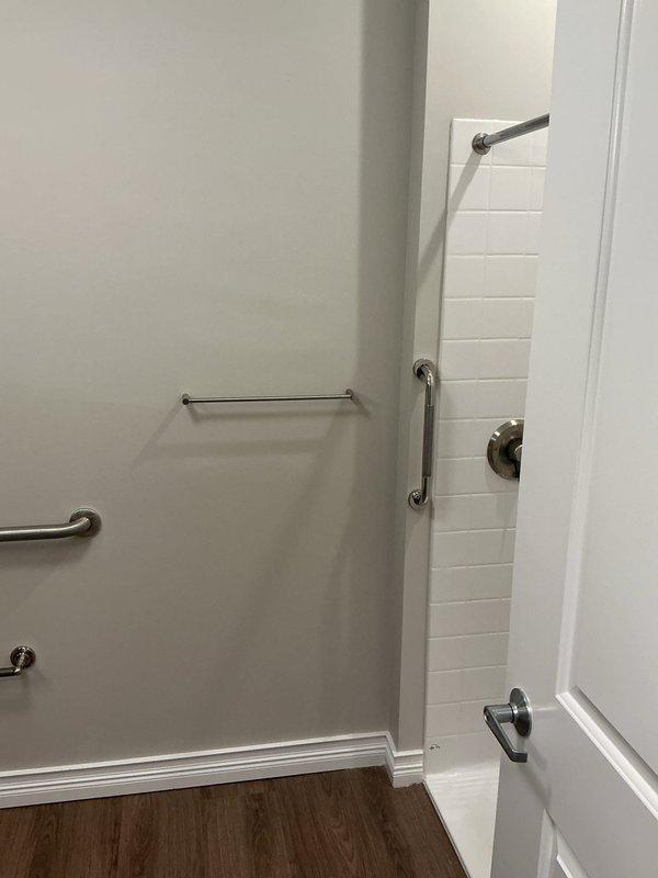 Evaluated existing shower supply line configuration and assessed feasibility of relocating water lines from attic space to accommodate revised bathroom layout per new construction plan. Inspected accessible bathroom installation featuring white subway tile surround, thermostatic mixing valve, handheld shower wand with slide bar, fixed showerhead, and multiple ADA-compliant grab bars in brushed nickel finish.