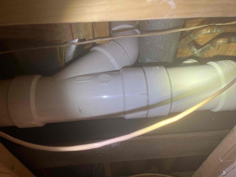 Completed return visit to finalize plumbing repair installation previously sold. Installed white PVC drainage piping system beneath subfloor with proper angular connections, elbow fittings, and directional changes secured to floor joists using plastic mounting straps. All pipe segments properly joined and clamped to wooden framing members to ensure stable, code-compliant installation. Documented completed work showing clean PVC drainage system with no visible leaks or defects, along with adjacent copper supply lines and mechanical components in crawl space area.