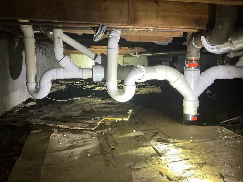 Completed comprehensive plumbing inspection and documentation of crawl space drainage system beneath residential structure. Inspected and photographed existing PVC drain line configuration including multiple P-trap assemblies, horizontal drain runs, and connection points to verify proper installation and identify any repairs needed per previous service agreement. All plumbing components, structural supports, and foundation access areas were examined and documented for service records.