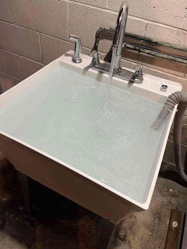 Responded to emergency call for utility sink collapse and major water leak causing flooding in basement area. Upon arrival, found white porcelain utility sink had detached from wall mounting, resulting in active water discharge onto concrete floor. Immediately shut off water supply to prevent further flooding damage.

Assessed structural damage to sink mounting system and determined cabinet support beneath sink had failed. Removed compromised sink assembly and inspected wall-mounted plumbing connections. Found copper supply lines and drainage system intact but required reconnection. Cleared standing water from work area and prepared surface for reinstallation.

Reinstalled utility sink with reinforced wall mounting brackets to ensure proper weight distribution and prevent future collapse. Secured cabinet base support structure and verified level positioning. Reconnected hot and cold water supply lines to chrome faucet assembly with pull-down spray head. Tested water pressure and inspec