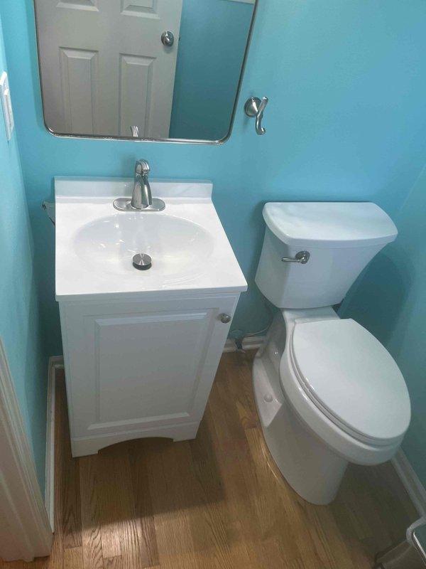 Returned to property to complete installation of customer-supplied bathroom vanity in small powder room with turquoise walls. Vanity unit consists of white cabinet base with integrated countertop and rectangular sink basin. Installed modern brushed nickel single-handle faucet with proper supply line connections and verified drain assembly with visible stopper mechanism functioning correctly. Secured vanity to wall studs, ensuring level placement on existing light wood-grain laminate flooring. Tested all plumbing connections for leaks, confirmed proper water flow and drainage. Mounted frameless rectangular mirror above vanity per standard placement. All fixtures operational and installation complete.