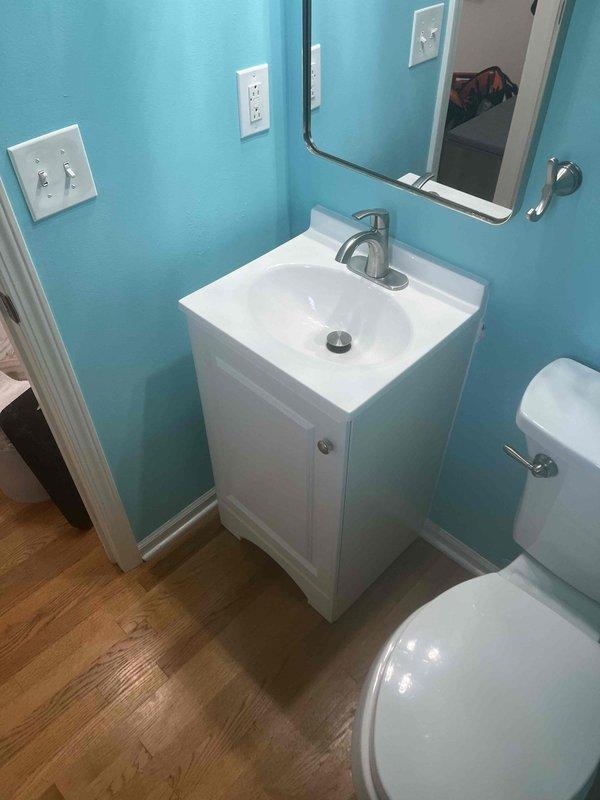 Returned to property to complete installation of customer-supplied bathroom vanity in small powder room with turquoise walls. Vanity unit consists of white cabinet base with integrated countertop and rectangular sink basin. Installed modern brushed nickel single-handle faucet with proper supply line connections and verified drain assembly with visible stopper mechanism functioning correctly. Secured vanity to wall studs, ensuring level placement on existing light wood-grain laminate flooring. Tested all plumbing connections for leaks, confirmed proper water flow and drainage. Mounted frameless rectangular mirror above vanity per standard placement. All fixtures operational and installation complete.