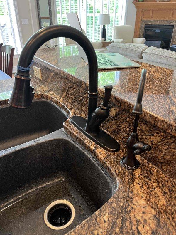 Completed replacement of two faucets in the residence: a chrome two-handle bathroom faucet with lever-style handles and lift rod assembly installed on a white ceramic sink, and an oil-rubbed bronze single-handle kitchen faucet with matching soap dispenser installed on granite countertop above an undermount sink. Both fixtures were properly mounted, connected to supply lines, tested for proper operation and leaks, with all components functioning correctly upon completion.