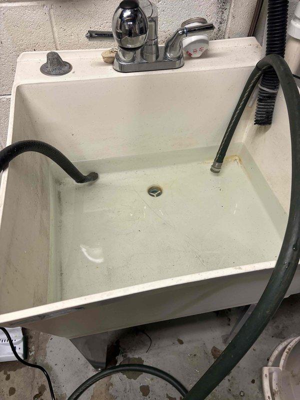 Responded to callback regarding exterior hose bibs dispensing hot water and persistent cloudy water condition one month after new water treatment system installation. Upon inspection, discovered cross-connection issue affecting outdoor faucets, causing hot water supply to both exterior fixtures. Investigated utility sink plumbing assembly where corroded galvanized piping beneath basin was identified as contributing factor to water discoloration. The heavily rusted metal pipes showed significant oxidation that could be introducing sediment into the water system. Assessed P-trap assembly and drainage components above the corroded section. Documented condition of existing plumbing infrastructure showing deteriorated metal piping requiring replacement to resolve ongoing water quality issues. Advised that foggy water appearance after one month indicates either inadequate system flushing following installation or continued contamination from degraded pipe interiors. Recommended complete repl