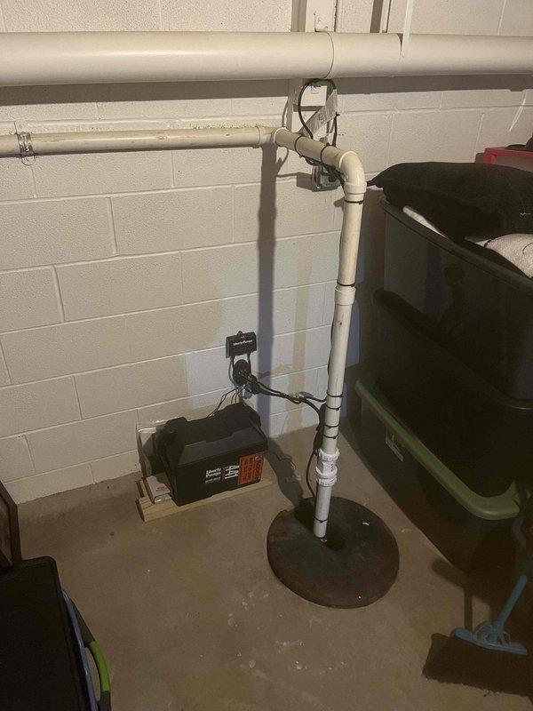Completed follow-up inspection on previously installed sump pump backup battery system. Verified proper installation of Liberty Pumps control unit mounted to foundation wall with secure electrical connections to PRO SERIES maintenance-free backup battery. Confirmed all wiring connections between control unit and battery terminals are tight and properly configured. Inspected vertical PVC discharge pipe assembly running from floor level through ceiling, ensuring all pipe sections are properly joined and secured with wall-mounted brackets. Verified floor flange installation at pipe base is secure and sealed. System tested and operating as designed to provide reliable backup power during primary pump or power failures.