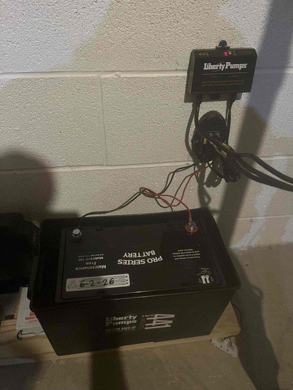Completed follow-up inspection on previously installed sump pump backup battery system. Verified proper installation of Liberty Pumps control unit mounted to foundation wall with secure electrical connections to PRO SERIES maintenance-free backup battery. Confirmed all wiring connections between control unit and battery terminals are tight and properly configured. Inspected vertical PVC discharge pipe assembly running from floor level through ceiling, ensuring all pipe sections are properly joined and secured with wall-mounted brackets. Verified floor flange installation at pipe base is secure and sealed. System tested and operating as designed to provide reliable backup power during primary pump or power failures.