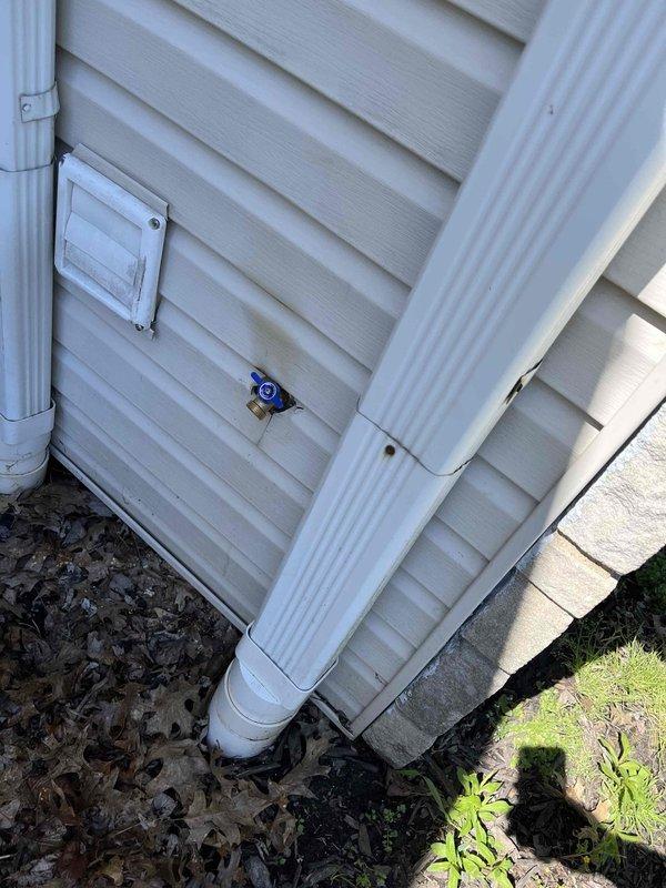 Installed outdoor hose bib with blue handle on exterior wall between two downspouts as part of promotional buy-one-get-one spigot offer. Completed installation on white vinyl siding with proper mounting and verified operational functionality of the new water spigot.