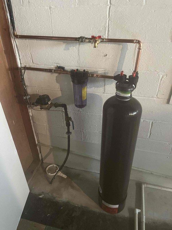 Completed installation of comprehensive whole house water treatment system in residential utility room. System includes tall cylindrical water softener with digital control head, inline sediment/carbon filtration housing with replaceable cartridge, and integrated copper piping manifold with brass ball valves and shutoff controls. All components professionally mounted to concrete block wall with proper drain line routing to floor drain. System fully pressurized, tested for leaks, and operational.
