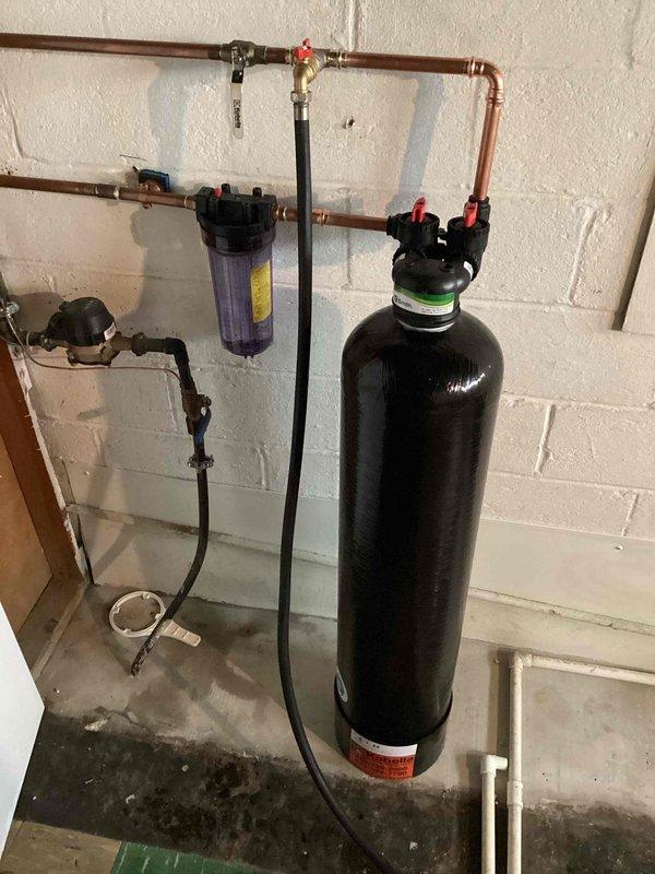 Completed installation of comprehensive whole house water treatment system in residential utility room. System includes tall cylindrical water softener with digital control head, inline sediment/carbon filtration housing with replaceable cartridge, and integrated copper piping manifold with brass ball valves and shutoff controls. All components professionally mounted to concrete block wall with proper drain line routing to floor drain. System fully pressurized, tested for leaks, and operational.