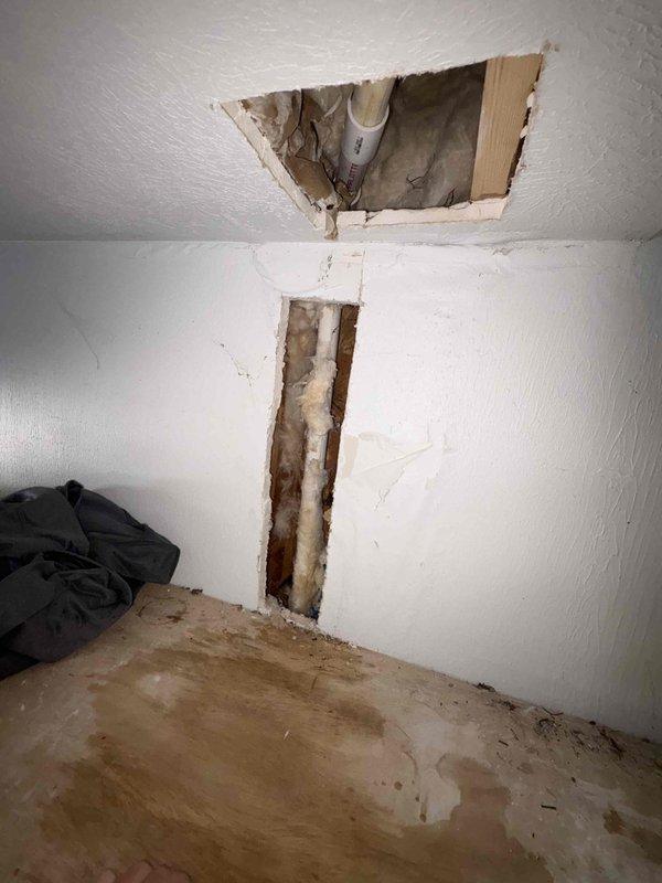 Returned to property to reconnect furnace condensate drain line to original configuration following previous water damage incident. Accessed repair area through existing drywall openings in wall and ceiling that exposed internal structure and PVC drainage system. Reassembled white PVC condensate drain piping using elbow fittings to restore proper drainage path from furnace unit. Performed operational test by running water through drain line to verify proper flow and confirm system functionality with no leaks or blockages detected. Evidence of previous water damage and moisture infiltration visible on wooden framing and floor surfaces in affected area.