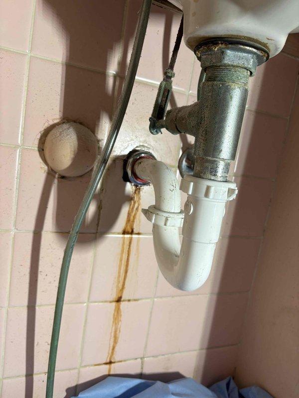 Completed repipe from kitchen sink to main stack and replaced lavatory drain line to main stack. Removed corroded metal drain fittings and deteriorated piping that showed evidence of long-term water leakage, as indicated by brown staining on surrounding tile surfaces. Installed new PVC drain assembly including P-trap and connecting pipes to eliminate leaks and restore proper drainage function. Work required access through cabinet area and attic/crawl space to route new piping to main plumbing stack.