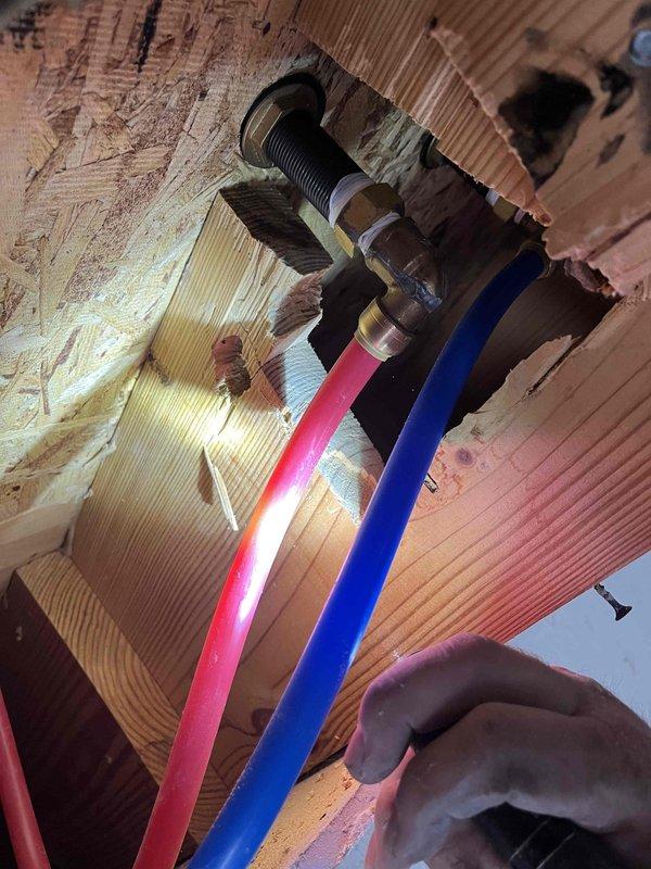 Responded to plumbing leak at supply line connection. Accessed piping through existing ceiling access panel using ladder. Located leak at threaded valve assembly where PEX supply lines (hot and cold) penetrate floor joists. Inspected flexible plastic supply lines and black threaded fitting mounted through subfloor. Repaired leaking connection at valve assembly to resolve recurring leak issue. Verified proper connection of pink hot water line and blue cold water line at penetration point. Restored system to proper working condition.