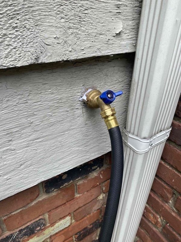 Installed new pre-meter main water shutoff valve system on building exterior using freeze machine technique to isolate water line without full system shutdown. Replaced existing shutoff with quarter-turn ball valve featuring blue handle for improved accessibility and operation, mounted to gray lap siding above brick foundation with brass fittings and flexible black connector hose for proper water flow control.