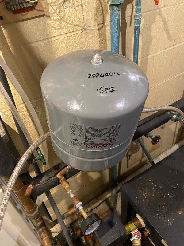 Returned to property to complete installation of pressure relief valve and expansion tank on hydronic heating system. Removed old expansion tank and associated fittings, then mounted new Amtrol expansion tank on existing wooden supports. Connected tank to system using copper piping and brass fittings, ensuring proper orientation and secure mounting. Installed new pressure relief valve with appropriate threaded connections to system piping. Verified all connections were tight and leak-free, checked system pressure on gauge, and confirmed proper operation of relief valve and expansion tank integration with existing circulator pump and controls.