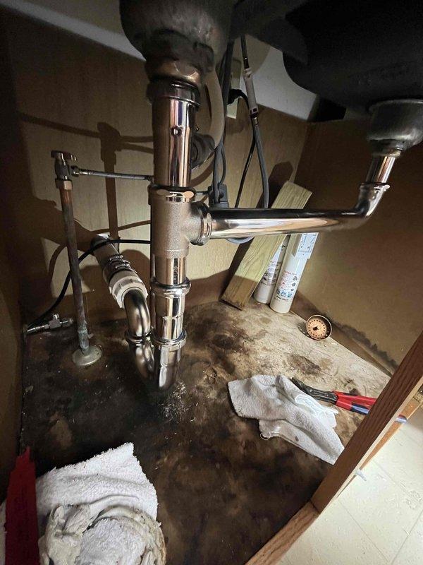 Responded to kitchen sink leak beneath cabinet. Upon inspection, found extensive water damage to cabinet floor with significant staining and deterioration from prolonged moisture exposure. Assessed plumbing assembly including garbage disposal unit, P-trap, and supply lines. Replaced deteriorated chrome piping and connections to eliminate active leak source. Cleaned and dried affected area, placing absorbent materials to address residual moisture. Tested all connections under pressure to verify proper seal and confirm leak resolution.