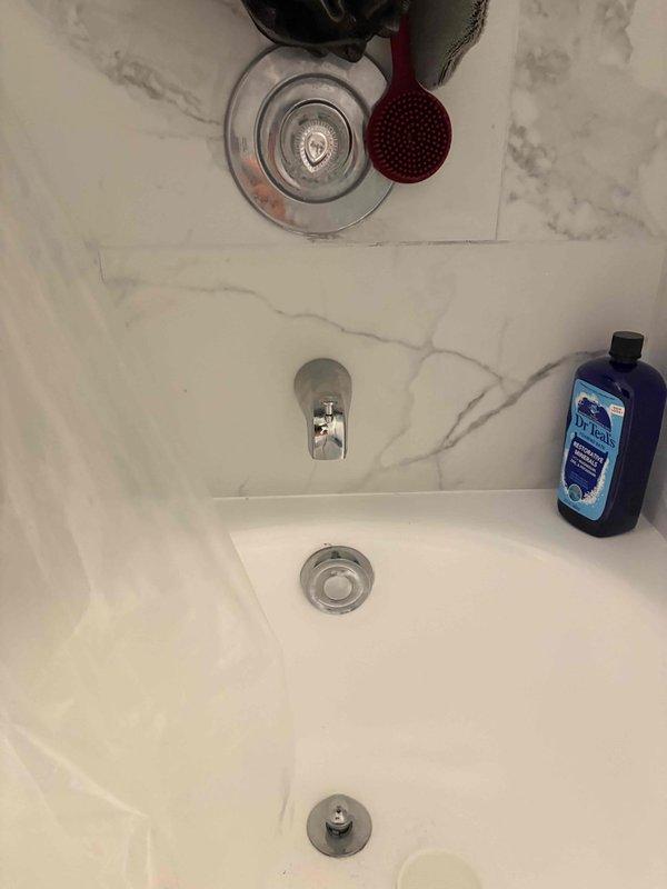 Responded to master bathroom bathtub drain leak in 1970s-era condominium. Performed comprehensive assessment of drain assembly, overflow plate, and spout connections. Took measurements of existing tub configuration and documented current fixture placement to evaluate repair feasibility versus potential walk-in shower conversion. Inspected underside access points and determined extent of work required, noting that drain repair may necessitate floor removal due to limited access in condo construction. Photographed existing conditions including chrome drain cover, overflow assembly, and surrounding marble-pattern tile work for warranty review and future reference.