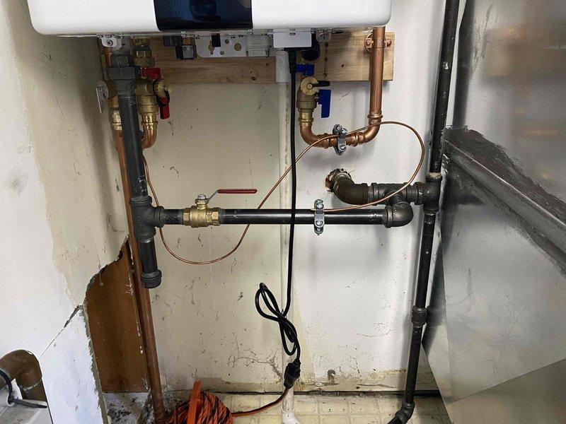 Completed sold repair work in residential laundry and water heater areas. Installed and connected dual water heater system with copper and black steel piping, including multiple shut-off valves with color-coded handles for system control. Configured plumbing connections for washing machine installation with exposed copper supply lines and drainage fittings on tile flooring.
