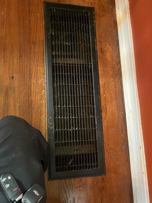 Conducted indoor air quality evaluation of existing floor-mounted cold air return system, estimated to be 60-70 years old. Performed inspection of ductwork infrastructure including crawl space access to assess condition of wooden structural components and measure dimensions for potential metal sheet lining installation. Documented existing floor register condition and surrounding wood flooring, noting age-related wear patterns around vent opening.