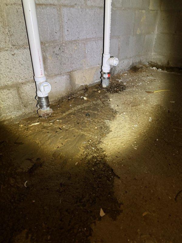 Responded to water leak at main water line entry point into residence. Upon arrival, found active leak at pipe penetration through foundation with no accessible shut-off valve at building entry. Main water shut-off valve and meter located 250 feet from structure in underground vault. Successfully located and accessed vault, shut off water supply to building to stop active leak and prevent further water damage to basement area. Basement floor showed significant water accumulation and moisture damage around pipe entry point where two white PVC vertical pipes penetrate concrete floor with metallic threaded transition fittings. Standing water and extensive floor staining indicated leak had been active for extended period. Photographically documented wet basement conditions, water damage patterns, pipe configurations, and deteriorated concrete surfaces for service records. Water service secured and isolated at vault location to allow for future repair work at building entry point.