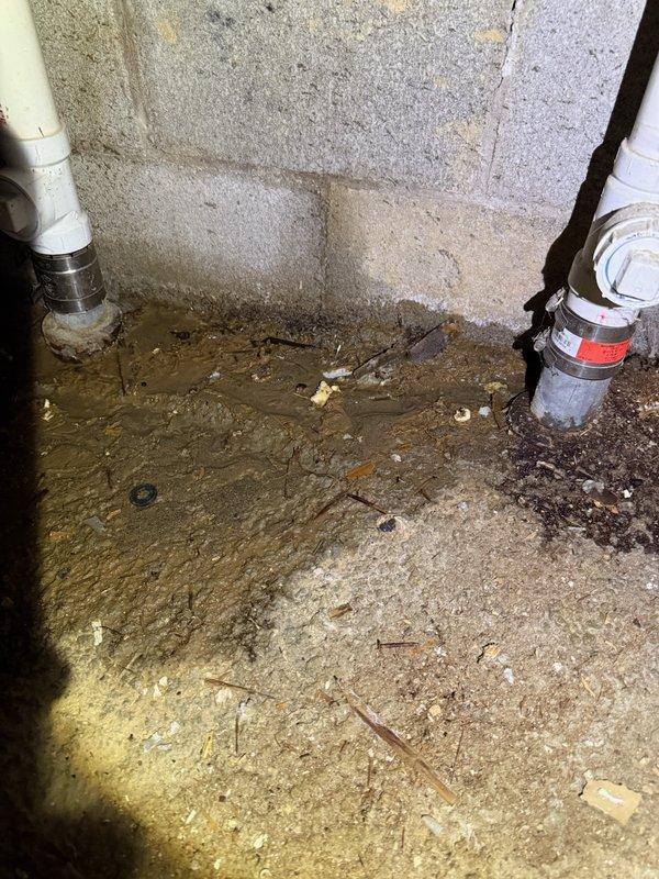Responded to water leak at main water line entry point into residence. Upon arrival, found active leak at pipe penetration through foundation with no accessible shut-off valve at building entry. Main water shut-off valve and meter located 250 feet from structure in underground vault. Successfully located and accessed vault, shut off water supply to building to stop active leak and prevent further water damage to basement area. Basement floor showed significant water accumulation and moisture damage around pipe entry point where two white PVC vertical pipes penetrate concrete floor with metallic threaded transition fittings. Standing water and extensive floor staining indicated leak had been active for extended period. Photographically documented wet basement conditions, water damage patterns, pipe configurations, and deteriorated concrete surfaces for service records. Water service secured and isolated at vault location to allow for future repair work at building entry point.
