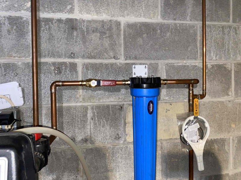 Returned to site to complete installation of new 40-gallon indirect water heater system with two technicians working full day on project. Integrated water heater into existing plumbing infrastructure with new copper piping, compression fittings, and brass connections mounted to basement wall. Installed inline water filtration system with blue housing and black cap, incorporating flow control valve and necessary shut-off components. Routed utility lines through basement ceiling joists, securing black flexible conduit, blue piping, and insulated lines with appropriate fasteners and zip ties. All connections tested and system confirmed operational upon completion of installation.