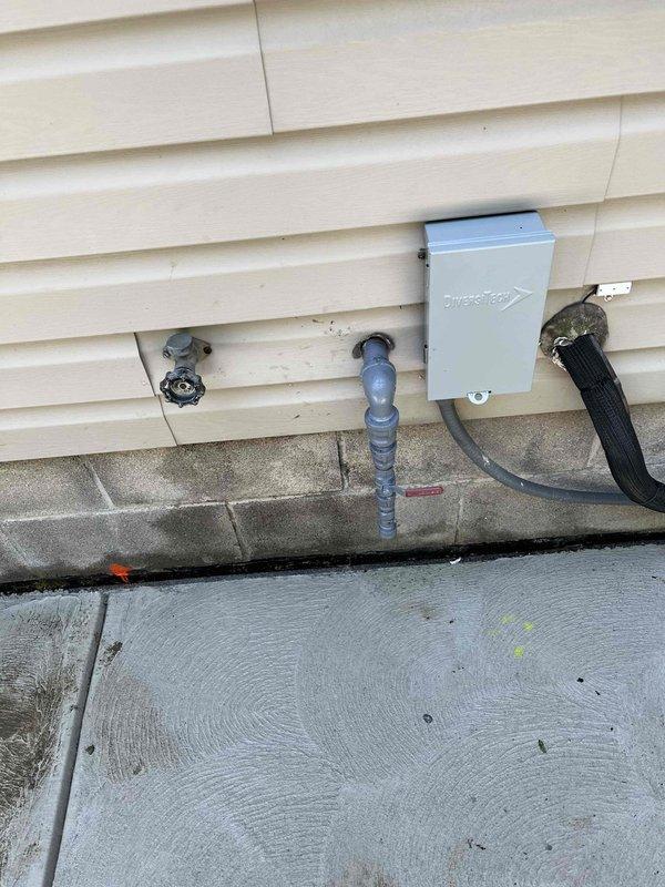 Serviced sump pump system experiencing heavy sludge accumulation. Inspected exterior discharge piping and verified proper drainage flow through outdoor discharge assembly. Documented existing installation showing weathered galvanized piping with blue valve control and PVC discharge line routing along building foundation.