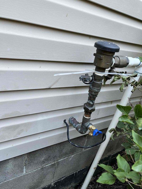 Serviced sump pump system experiencing heavy sludge accumulation. Inspected exterior discharge piping and verified proper drainage flow through outdoor discharge assembly. Documented existing installation showing weathered galvanized piping with blue valve control and PVC discharge line routing along building foundation.