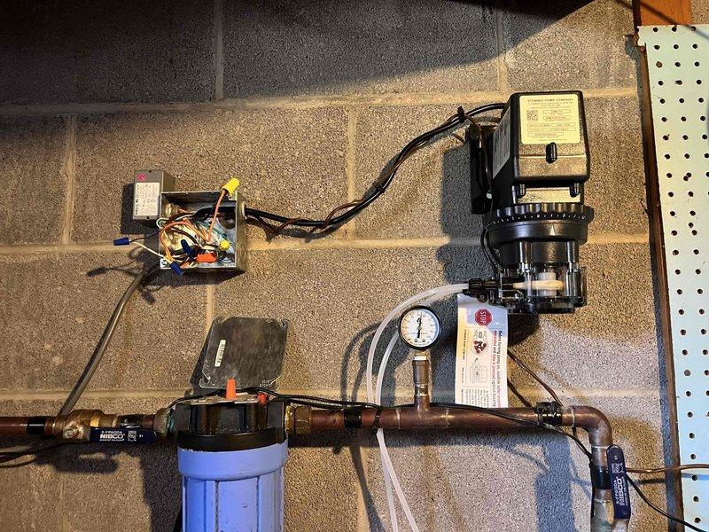 Responded to service call for non-operational Stenner chemical feed pump with no power indicator light. Upon arrival, performed diagnostic testing using digital multimeter to check electrical components and circuit board functionality. Removed pump housing cover to access internal electronics and identified power supply issue. Tested voltage levels at various connection points on the green circuit board to isolate the electrical fault. Inspected the complete water treatment system installation including blue filter housing, copper piping, pressure gauge, and associated electrical control components to ensure proper integration with the chemical feed pump. Verified all plumbing connections and electrical wiring from the control box to the pump unit were secure and properly terminated. Completed troubleshooting and testing procedures to restore pump operation and system functionality.