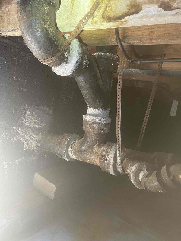 # Clogged Drain/Sink Service

Responded to service call for clogged sink and bathtub at residential property. Upon inspection, accessed crawl space to examine drainage system and identified severely deteriorated cast iron drain piping with heavy corrosion, rust buildup, and mineral deposits throughout the system. The horizontal drain lines showed pronounced scaling and surface degradation, with visible efflorescence around pipe joints indicating long-term moisture issues and compromised pipe integrity. Vertical drain stacks displayed similar weathering and discoloration consistent with aging cast iron plumbing infrastructure.

Documented existing conditions of the drainage system, including corroded pipe sections suspended from floor joists with metal hanger straps. The deteriorated state of the piping likely contributing to reduced flow capacity and drainage issues reported by customer. Photographed the affected plumbing components and structural configuration for service records and 