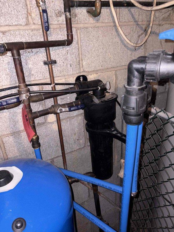 Conducted site evaluation of existing plumbing and water treatment system to revisit previous year's replacement estimates. Documented current configuration including water softener with blue pressure tank, whole-house filtration system with black cylindrical housing, copper and PEX piping distribution network, multiple shut-off valves with red gate handles, and aging brass fittings showing oxidation. Assessed condition of mixed-material installation featuring copper pipes in various stages of wear, plastic components, and mounted equipment on concrete block and wooden framing to prepare updated replacement recommendations.