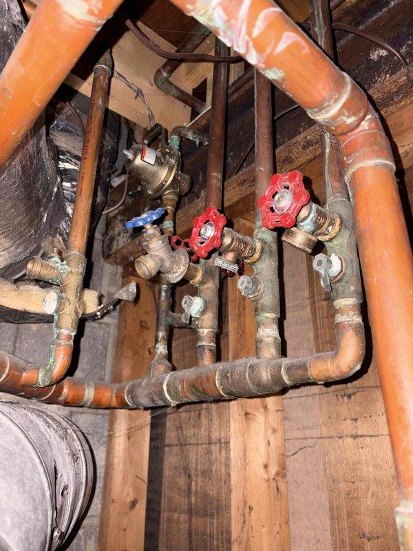 Conducted site evaluation of existing plumbing and water treatment system to revisit previous year's replacement estimates. Documented current configuration including water softener with blue pressure tank, whole-house filtration system with black cylindrical housing, copper and PEX piping distribution network, multiple shut-off valves with red gate handles, and aging brass fittings showing oxidation. Assessed condition of mixed-material installation featuring copper pipes in various stages of wear, plastic components, and mounted equipment on concrete block and wooden framing to prepare updated replacement recommendations.