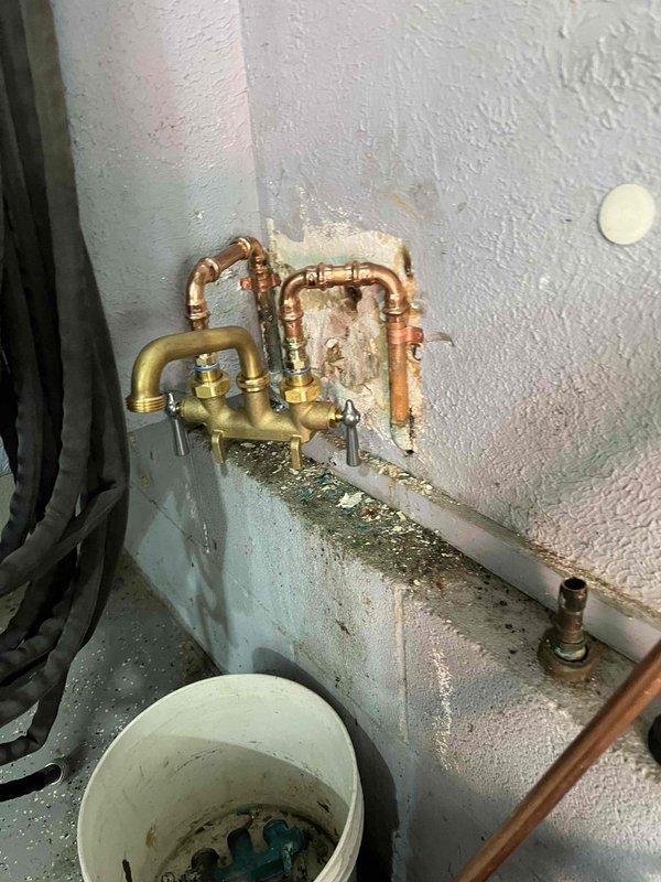 # Plumbing Service Completed

Returned to property to complete hose bib installation that was previously sold and initiated. Upon arrival, assessed existing plumbing configuration featuring aged copper piping with brass manifold connections mounted to exterior wall penetration area. Wall surface showed significant deterioration and damage around pipe penetrations, with exposed substrate material and moisture intrusion evident throughout the work area.

Completed installation of hose bib assembly, making necessary connections to existing copper supply lines through brass manifold system. All pipe connections were properly secured using appropriate fittings and clamps to ensure water-tight seals. Verified proper valve operation and flow control functionality of newly installed hose bib.

Addressed moisture concerns by inspecting all visible piping for leaks and confirming integrity of connections. Noted heavy condensation on wall surfaces and piping components, along with mineral deposit