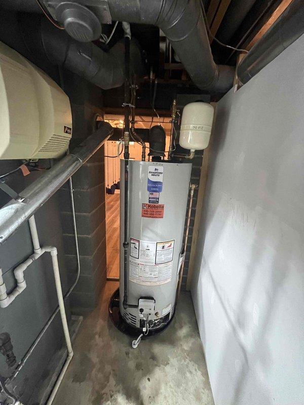 Responded to reported hot water tank leak at 1970s-era residential property. Upon arrival, conducted thorough inspection of Bradford White water heater unit and surrounding plumbing infrastructure. Assessed visible leak source, examined tank condition, connections, and associated copper piping system. Verified operational status of heating elements through inspection port showing active combustion. Documented existing installation configuration including expansion tank, PVC drainage system, and copper supply lines. Photographed equipment condition and mechanical room setup for service records. Identified leak origin point and evaluated overall system integrity. Provided assessment findings and recommended appropriate repair or replacement options based on unit age and condition of 50-year-old home's plumbing system.