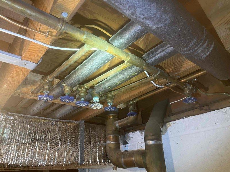Completed plumbing estimate for basement piping replacement project. Assessed multiple corroded shut-off valves with blue handles installed on copper water supply lines beneath floor joists, noting extensive condensation on existing foam pipe insulation throughout the system. Evaluated deteriorated S-traps and sink drain assemblies requiring replacement, including chrome-finished P-trap configuration with vertical drain connections and braided stainless steel supply lines mounted to wall surface. Documented current conditions of plumbing fixtures, pipe fittings, and drainage components to provide accurate replacement recommendations for corroded valves and trap assemblies.
