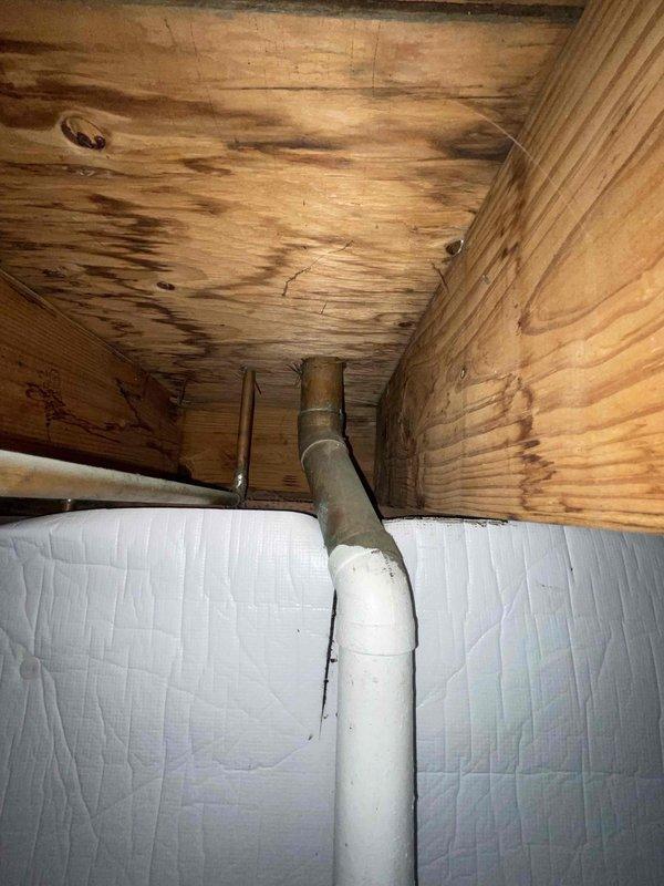 Responded to water leak complaint in basement area beneath kitchen sink. Upon inspection, discovered active leak originating from corroded metal drain pipe section. The deteriorated pipe showed advanced rust and oxidation, with heavy corrosion causing water seepage onto concrete floor and surrounding wooden cabinet structure. Water staining was evident on wooden ceiling panels inside cabinet, indicating ongoing moisture intrusion. Identified transition point between white PVC upper drain line and corroded metal lower section as primary failure zone. The metal pipe exhibited severe degradation with flaking surfaces and rust buildup compromising pipe integrity. Documented damage with photographs showing extent of corrosion and water staining. Recommended immediate replacement of corroded metal pipe section with PVC to prevent further water damage and restore proper drainage function.