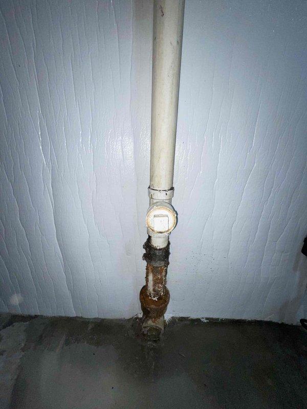 Responded to water leak complaint in basement area beneath kitchen sink. Upon inspection, discovered active leak originating from corroded metal drain pipe section. The deteriorated pipe showed advanced rust and oxidation, with heavy corrosion causing water seepage onto concrete floor and surrounding wooden cabinet structure. Water staining was evident on wooden ceiling panels inside cabinet, indicating ongoing moisture intrusion. Identified transition point between white PVC upper drain line and corroded metal lower section as primary failure zone. The metal pipe exhibited severe degradation with flaking surfaces and rust buildup compromising pipe integrity. Documented damage with photographs showing extent of corrosion and water staining. Recommended immediate replacement of corroded metal pipe section with PVC to prevent further water damage and restore proper drainage function.