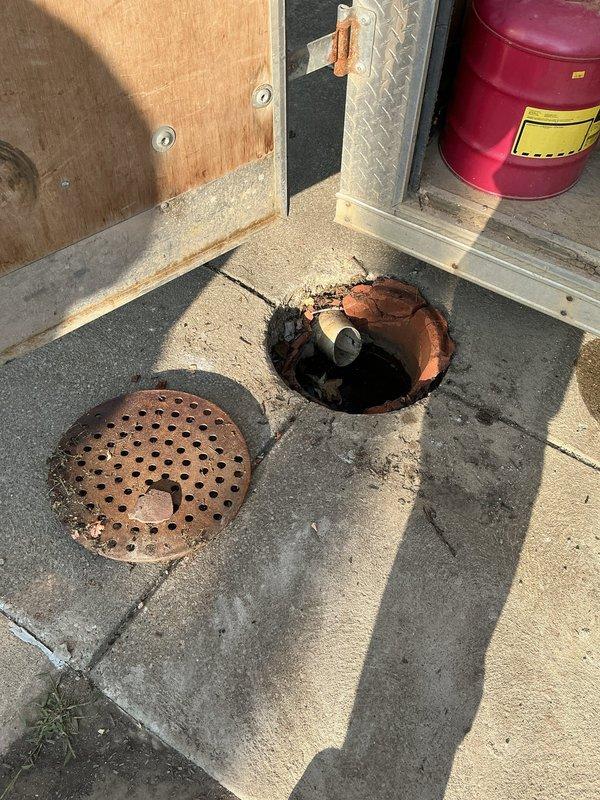 Conducted comprehensive assessment of deteriorated sewer drain crock system requiring replacement. Inspection revealed damaged terracotta clay pipe fragments and compromised subsurface infrastructure with significant pavement deterioration extending approximately 14-22 inches deep. Documented exposed PVC piping section showing angular displacement and interior residue accumulation. Secondary inspection identified corroded perforated metal drainage cover with extensive rust deterioration and open access point exposing interior plumbing components. Provided detailed photographic documentation of both street-level excavation site and interior floor drainage system to support replacement estimate for complete sewer drain crock assembly.