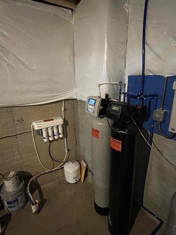 Returned to complete installation of comprehensive residential water treatment system in basement utility area. System consists of black cylindrical water softener with digital control head, gray pressure tank, wall-mounted white multi-stage filtration unit with multiple filter housings, and blue control box with electrical connections. Verified all plumbing connections including silver metallic pipes, white PVC piping, and associated fittings are secure and properly configured. Confirmed proper operation of water softener control panel, pressure tank mounting, and filtration system components. All equipment functioning as designed with appropriate warning labels visible and system ready for service.