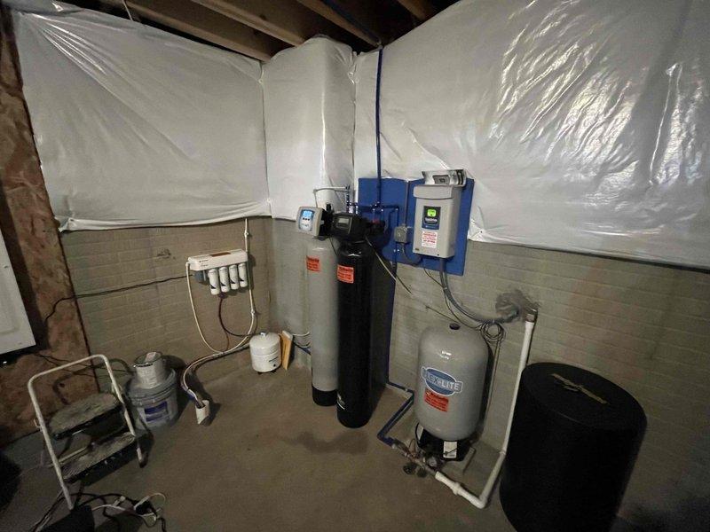 Returned to complete installation of comprehensive residential water treatment system in basement utility area. System consists of black cylindrical water softener with digital control head, gray pressure tank, wall-mounted white multi-stage filtration unit with multiple filter housings, and blue control box with electrical connections. Verified all plumbing connections including silver metallic pipes, white PVC piping, and associated fittings are secure and properly configured. Confirmed proper operation of water softener control panel, pressure tank mounting, and filtration system components. All equipment functioning as designed with appropriate warning labels visible and system ready for service.