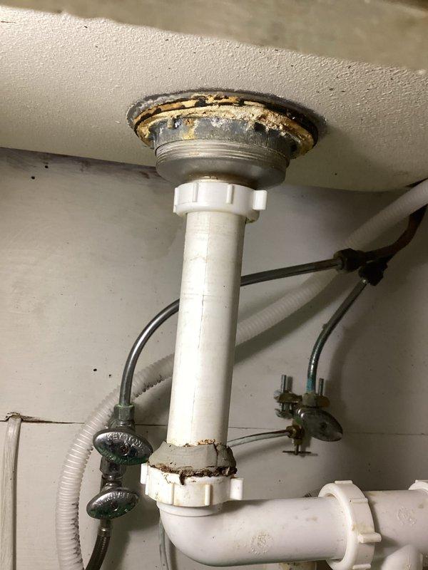 Completed plumbing membership inspection at residential property. Documented double-basin kitchen sink installation with chrome faucet and assessed under-sink plumbing components. Noted significant corrosion and deterioration on drain flange mounting assembly with visible rust and mineral deposits, along with aging P-trap and supply line connections requiring future attention.