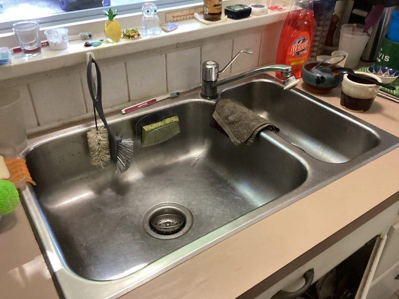 Completed plumbing membership inspection at residential property. Documented double-basin kitchen sink installation with chrome faucet and assessed under-sink plumbing components. Noted significant corrosion and deterioration on drain flange mounting assembly with visible rust and mineral deposits, along with aging P-trap and supply line connections requiring future attention.