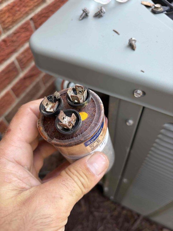 Responded to no cooling complaint with indoor air handler operating but outdoor unit fan not running. Upon inspection, found failed dual run capacitor with severely corroded terminals and rust damage. Removed defective capacitor and installed new replacement unit. Tested contactor operation and verified all electrical connections secure. System restored to normal operation with outdoor condenser fan and compressor running properly.