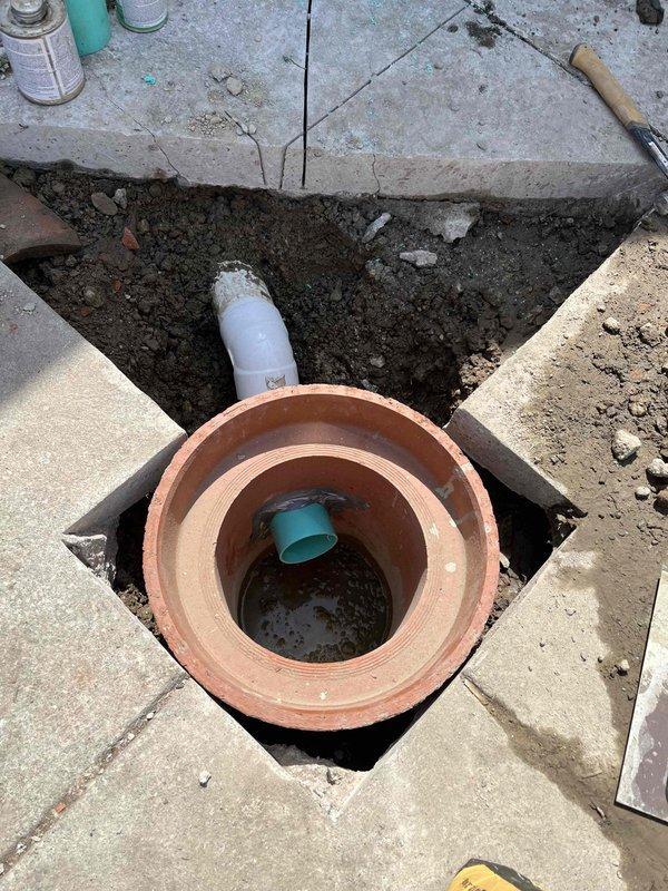 Returned to property to complete installation of new clay tile driveway crock as previously sold. Excavated existing deteriorated concrete surrounding old cast iron cleanout fitting. Removed corroded metal component and replaced with new terracotta clay tile crock fitting. Connected existing white PVC drainage pipe to new fitting, ensuring proper alignment and seal. Backfilled excavation area and prepared site for concrete restoration around new cleanout assembly.