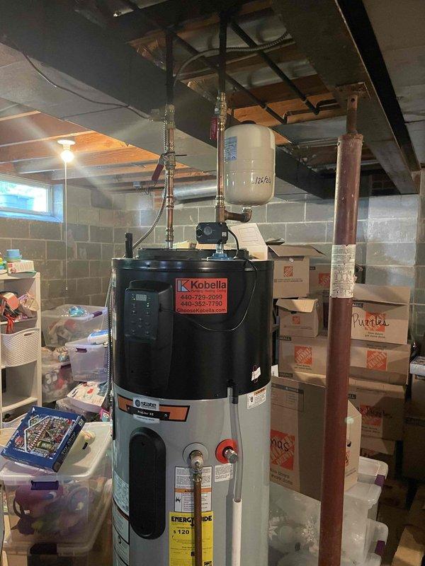Responded to complaint of hot water tank depleting within five minutes of use throughout the residence. Upon arrival, inspected Kobella brand tanked water heater that was recently installed in February in basement mechanical room. Verified proper installation of expansion tank with date marking on unit surface, copper supply and discharge piping with brass fittings and shut-off valves, and all connections secure without visible leaks. Examined water heater control panel settings and EnergyGuide specifications to confirm unit capacity appropriate for household demand. Assessed plumbing configuration including flexible braided connectors and valve positioning. Determined potential issue with thermostat calibration or heating element performance causing premature hot water depletion. Documented equipment condition and installation quality through photographic records showing expansion tank, piping system, and water heater unit in unfinished basement setting. Recommended monitoring water t
