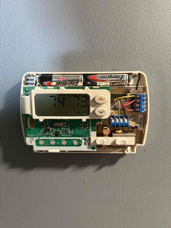 Conducted comprehensive HVAC system assessment for equipment replacement estimate. Documented existing thermostat configuration, revealing wall-mounted digital programmable unit with exposed internal components including circuit board, wiring terminals, and battery backup system. Evaluated exterior HVAC equipment location at building foundation, noting weathered concrete pad, utility connections, and existing refrigerant line routing along exterior wall. Assessment identified aging infrastructure requiring replacement to restore system efficiency and reliability.