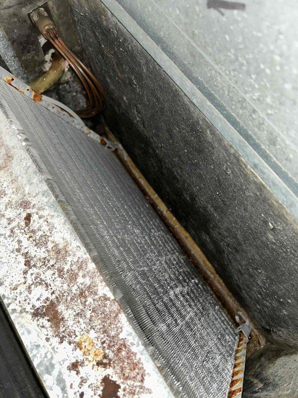 Completed cooling maintenance club visit on air handler system. Upon inspection, found evaporator coil with heavy dirt and mold accumulation on fins, requiring attention. Noted significant oxidation and corrosion on copper refrigerant lines with greenish-brown patina and rust staining. Observed rust formation and debris buildup on unit housing and frame. Documented condition of refrigerant line connections and insulated suction line passing through metal panel. System shows signs of extended operational use with maintenance needs identified for future service recommendations.